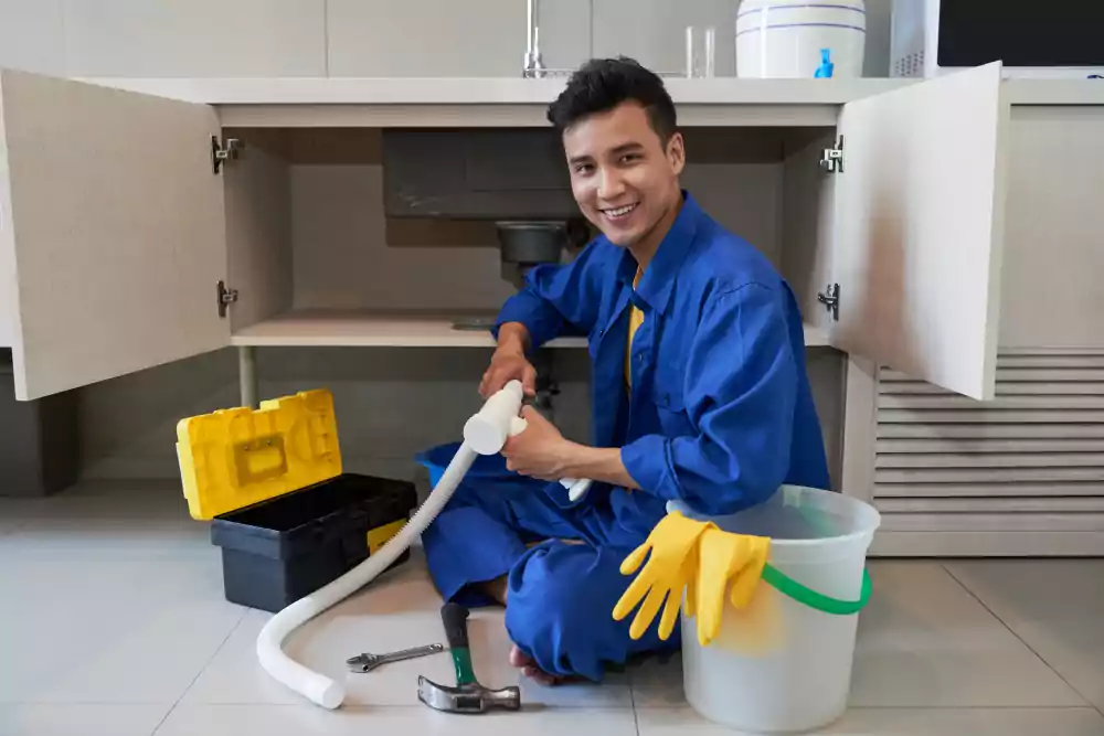 cheerful asian plumber sitting floor repairing kitchen sink